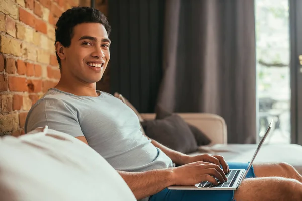 Uomo razza mista sorridente agghiacciante con computer portatile sul divano in quarantena — Foto stock