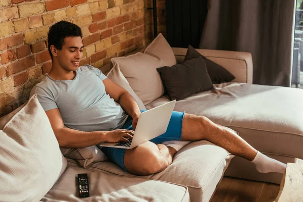 Sorrindo misto raça homem refrigeração com laptop no sofá com controle remoto durante o auto isolamento — Fotografia de Stock