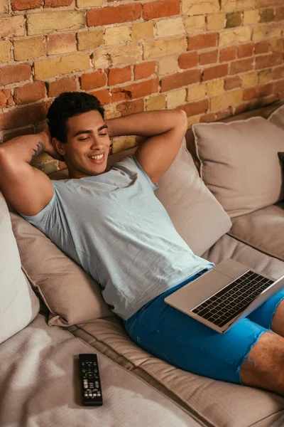 Gai homme bi-racial refroidissant avec ordinateur portable sur canapé avec télécommande pendant l'isolement personnel — Photo de stock