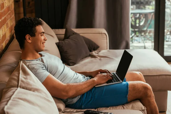 Homme de race mixte souriant utilisant un ordinateur portable avec écran vierge en quarantaine — Photo de stock