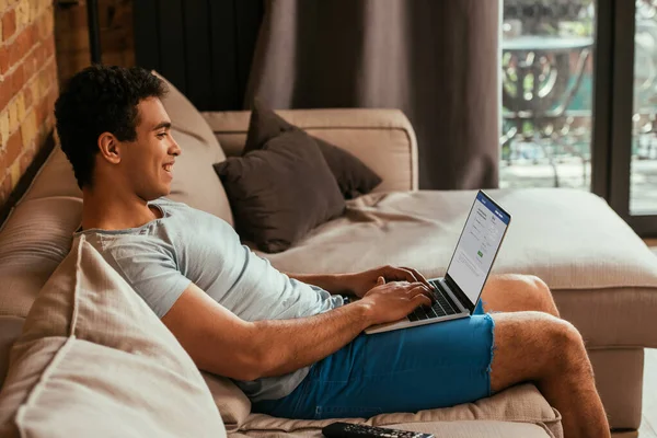 KYIV, UKRAINE - 27 de abril de 2020: homem de raça mista sorridente usando laptop com facebook na tela durante o auto-isolamento — Fotografia de Stock