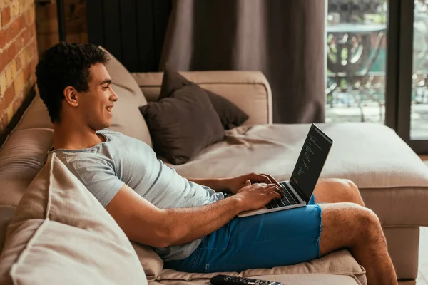 KYIV, UKRAINE - APRIL 27, 2020: happy mixed race man using laptop with html code on screen during self isolation — Stock Photo