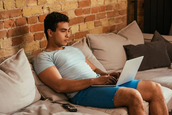 Jeune homme de race mixte travaillant sur un ordinateur portable sur un canapé pendant l'isolement personnel — Photo de stock