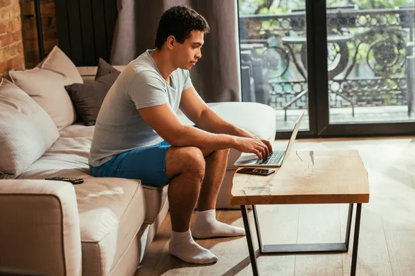 Giovane freelance di razza mista che lavora con laptop sul divano durante l'autoisolamento — Foto stock