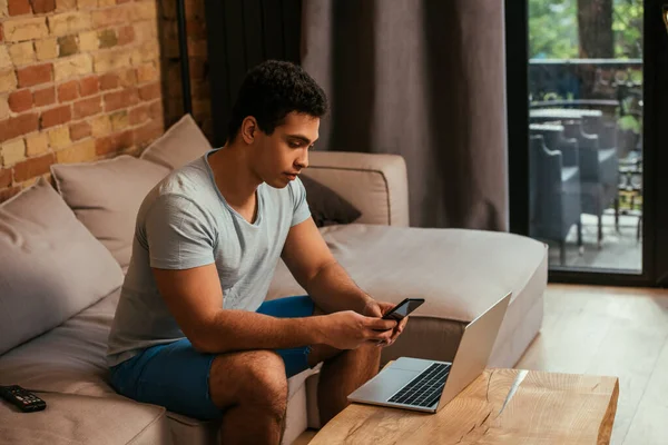 Gutaussehender Mixed Race Freelancer, der während der Selbstisolierung mit Smartphone und Laptop auf dem Sofa arbeitet — Stockfoto