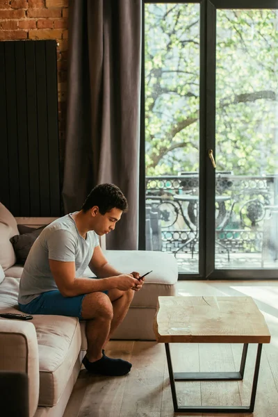 Giovane uomo di razza mista utilizzando smartphone sul divano durante la quarantena — Foto stock