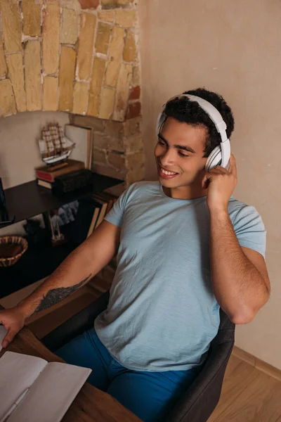 Sonriente freelancer de carrera mixta trabajando con auriculares y bloc de notas en la oficina en casa durante el autoaislamiento - foto de stock