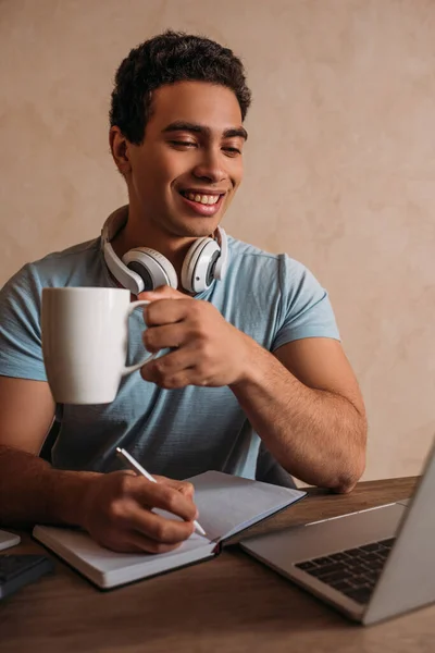 Sonriente carrera mixta freelancer sosteniendo taza de café mientras trabaja con el ordenador portátil y bloc de notas en la oficina en el hogar durante el autoaislamiento - foto de stock