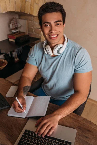 Усміхнений фрілансер змішаної раси, що працює з ноутбуком та блокнотом в домашньому офісі під час карантину — стокове фото