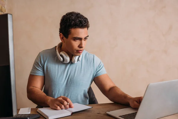 Apuesto freelancer de raza mixta que trabaja con el ordenador portátil, y bloc de notas en la oficina en el hogar durante el autoaislamiento - foto de stock