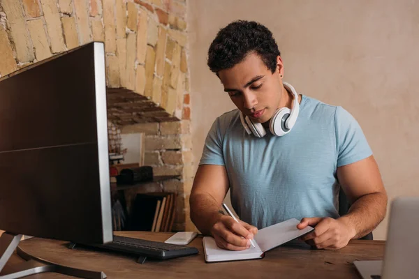 Freelancer de carrera mixta escribiendo en bloc de notas y trabajando en la oficina en casa durante el autoaislamiento - foto de stock