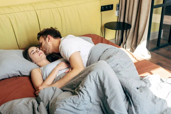 Bearded man kissing attractive girlfriend with closed eyes lying in bed — Stock Photo