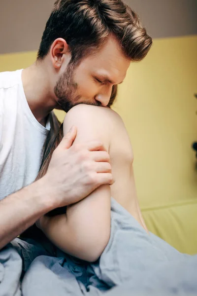 Selective focus of handsome man kissing shoulder of sensual girlfriend in bedroom — Stock Photo