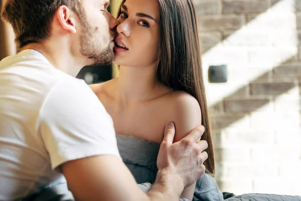 Selektiver Fokus eines bärtigen Mannes mit geschlossenen Augen, der eine attraktive und sinnliche Freundin im Schlafzimmer küsst — Stockfoto