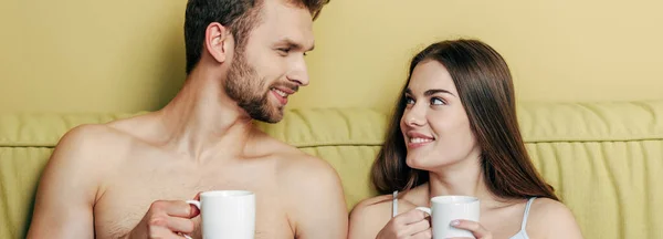 Imagen horizontal de pareja feliz mirándose y sosteniendo tazas con café - foto de stock