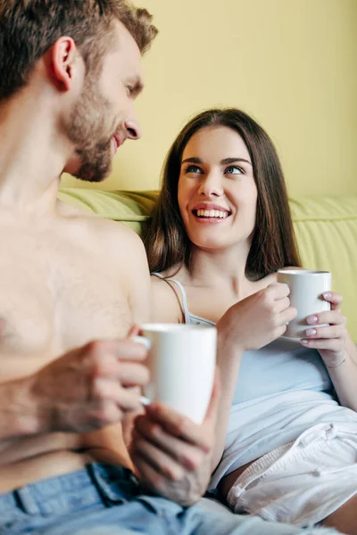 Enfoque selectivo de pareja alegre sosteniendo tazas con café y mirándose en el dormitorio - foto de stock