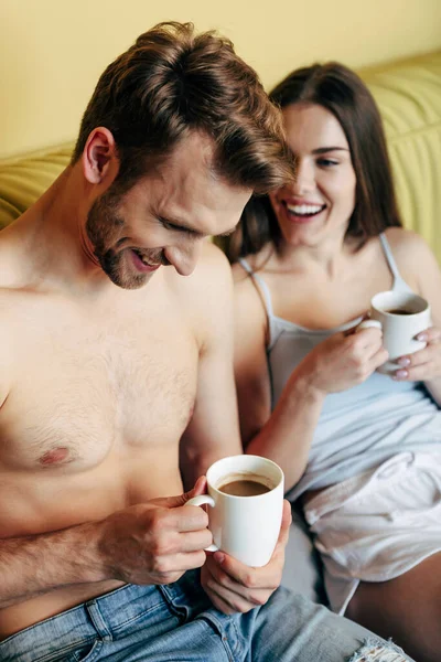 Selective focus of cheerful woman looking at muscular boyfriend and holding cup with coffee in bedroom — Stock Photo