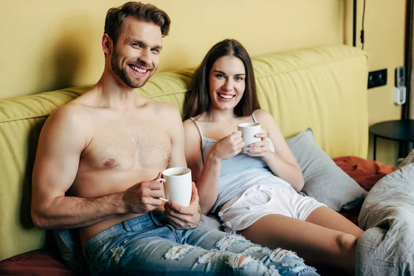 Feliz hombre musculoso y mujer alegre mirando a la cámara mientras sostiene tazas con café - foto de stock