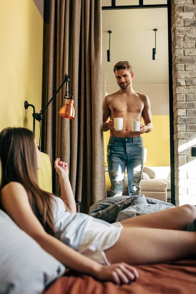 Enfoque selectivo de hombre muscular sosteniendo tazas cerca de la mujer en el dormitorio - foto de stock