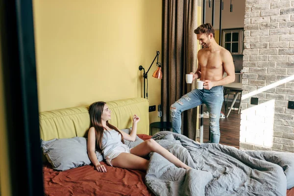 Enfoque selectivo del hombre musculoso sonriendo mientras sostiene tazas cerca de la mujer en el dormitorio - foto de stock