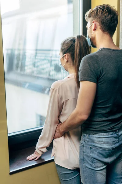 Мужчина обнимает девушку в медицинской маске и стоит у окна — стоковое фото