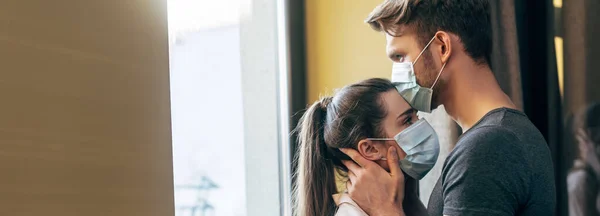 La cosecha panorámica del hombre en la máscara médica que besa la frente de la mujer en casa - foto de stock