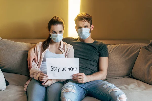 Uomo e donna in maschere mediche seduti sul divano e tenendo cartello con soggiorno a casa lettering — Foto stock