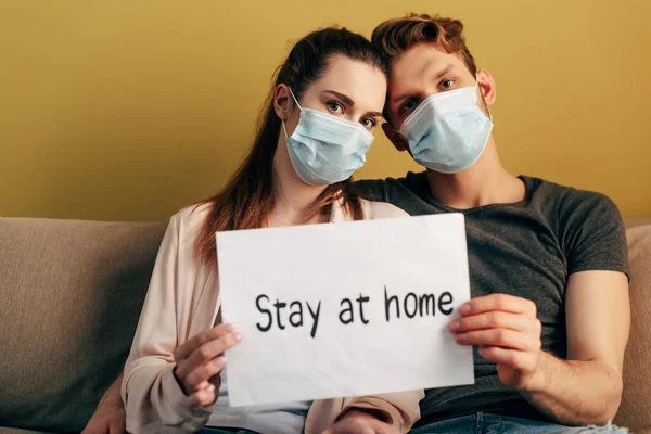 Selective focus of man and woman in medical masks sitting on sofa and holding placard with stay at home lettering — Stock Photo