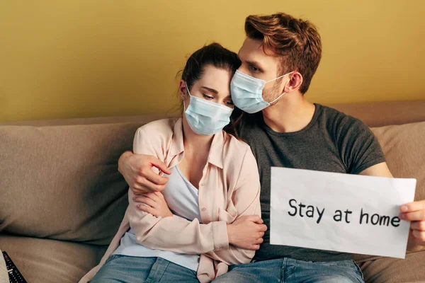 Selektiver Fokus des Mannes umarmt Freundin in medizinischer Maske und hält Plakat mit Bleibe-Schriftzug hoch — Stockfoto