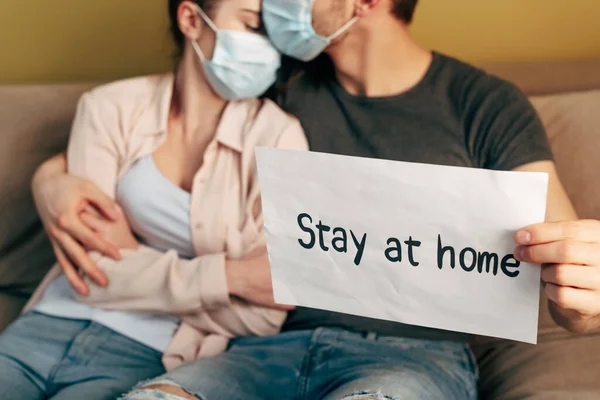 Messa a fuoco selettiva di uomo abbracciando ragazza in maschera medica e tenendo cartello con soggiorno lettering a casa — Foto stock
