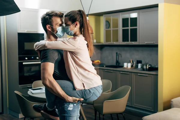 Hombre en máscara médica celebración en brazos chica en sala de estar moderna - foto de stock