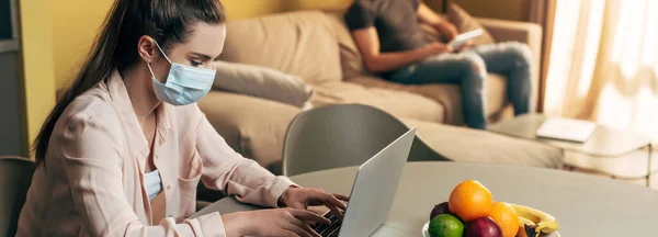 Панорамний знімок фрілансера в медичній масці з ноутбуком біля людини з цифровим планшетом вдома — стокове фото