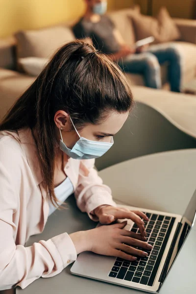 Enfoque selectivo de freelancer en máscara médica utilizando el ordenador portátil cerca del hombre en casa - foto de stock