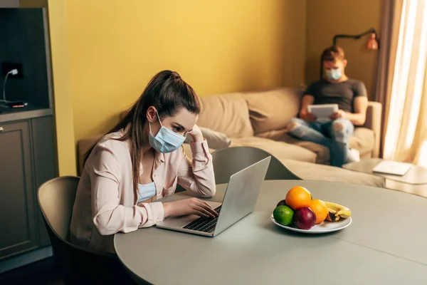 Foyer sélectif de pigiste dans le masque médical à l'aide d'un ordinateur portable près des fruits et l'homme avec tablette numérique à la maison — Photo de stock