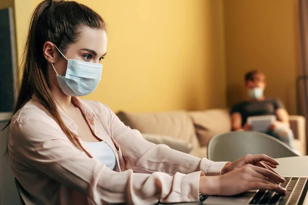 Enfoque selectivo de freelancer en la escritura de máscara médica en el ordenador portátil cerca del hombre en casa - foto de stock