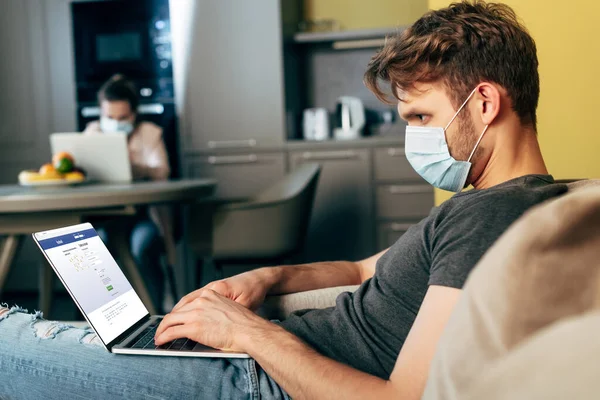 KYIV, UKRAINE - APRIL 22, 2020: selective focus of freelancer in medical mask using laptop with facebook website near girl — стокове фото