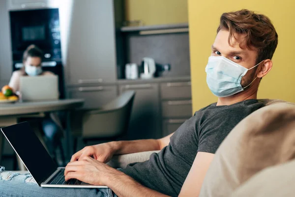 Foco seletivo do freelancer em máscara médica usando laptop com tela em branco perto da namorada — Fotografia de Stock