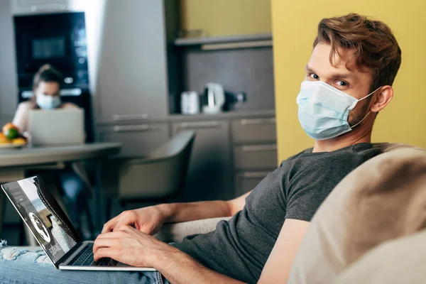 Selective focus of freelancer in medical mask using laptop with booking website near girlfriend — Stock Photo