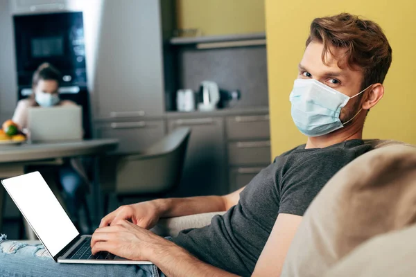 Selective focus of freelancer in medical mask using laptop with white screen near girlfriend — Stock Photo