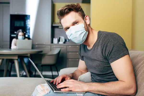 Selective focus of freelancer in medical mask near laptop and girlfriend at home — Stock Photo