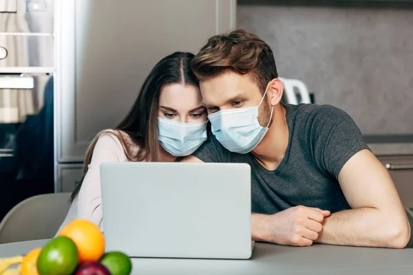 Selective focus of couple in medical masks watching movie on laptop near fruits — Stock Photo