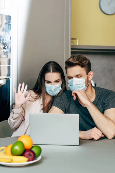 Selective focus of girl in medical mask waving hand while having video chat near man — Stock Photo