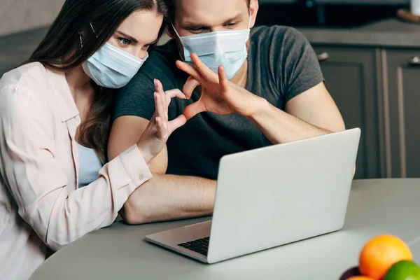 Selective focus of couple in medical masks showing heart with hands while having video chat — Stock Photo