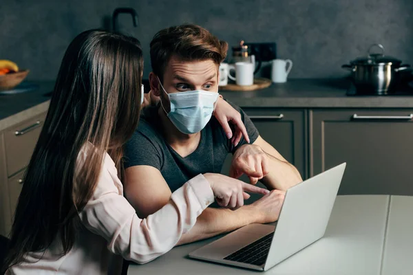 Mujer señalando con el dedo a la computadora portátil cerca de novio en máscara médica - foto de stock