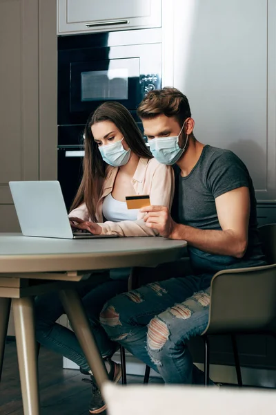 Enfoque selectivo del hombre en la máscara médica que sostiene la tarjeta de crédito mientras está sentado cerca del ordenador portátil y la niña - foto de stock