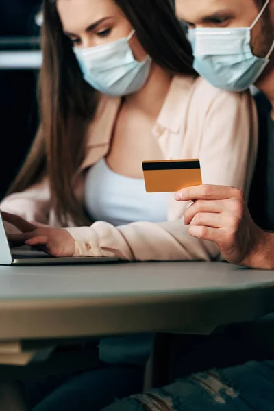 Enfoque selectivo del hombre en máscara médica celebración de la tarjeta de crédito, mientras que la chica de compras en línea - foto de stock