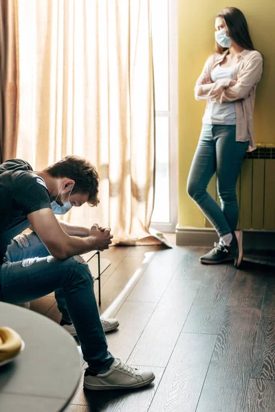 Selective focus of man in medical mask sitting near offended girlfriend with crossed arms — Stock Photo