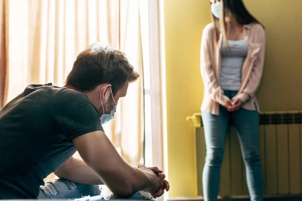 Foco seletivo do homem em máscara médica sentado com as mãos apertadas perto da namorada ofendida — Fotografia de Stock