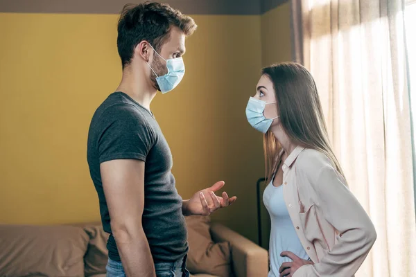 Visão lateral do homem em gestos máscara médica enquanto briga com a mulher em casa — Fotografia de Stock