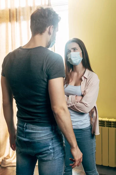 Donna in maschera medica in piedi con le braccia incrociate mentre litiga con il fidanzato a casa — Foto stock
